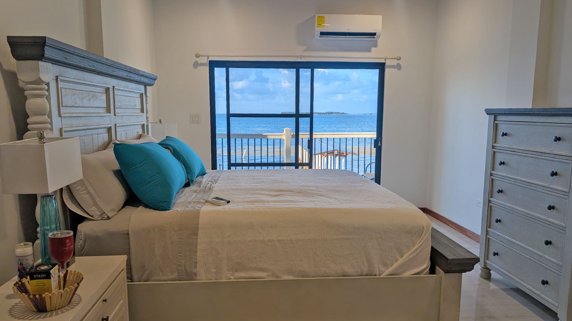 A cozy bedroom with a light-colored bed and turquoise pillows, featuring a view of the ocean through a large window.