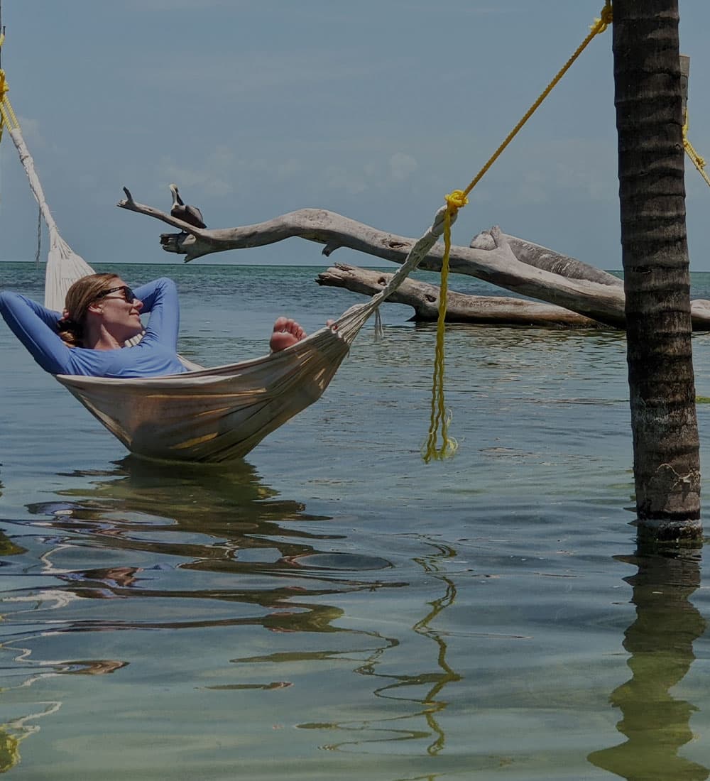 A person relaxes in a hammock over shallow water with a driftwood backdrop.